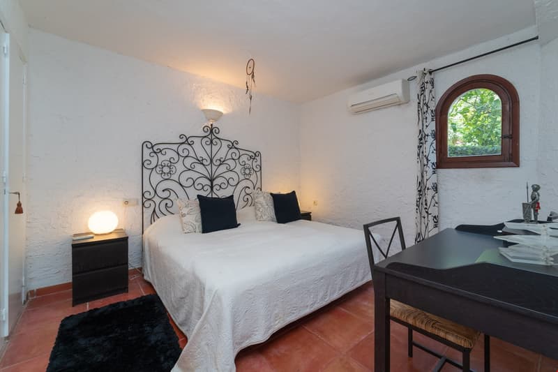 Bedroom with white walls, wrought iron headboard, white bedding, black and white patterned curtains, wooden desk, and a window overlooking greenery outside.