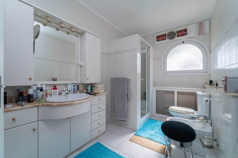 Spacious bathroom with a vanity sink, mirror, and storage cabinets. There is a toilet, a window, and a towel rack. The room has tiled floors and walls.