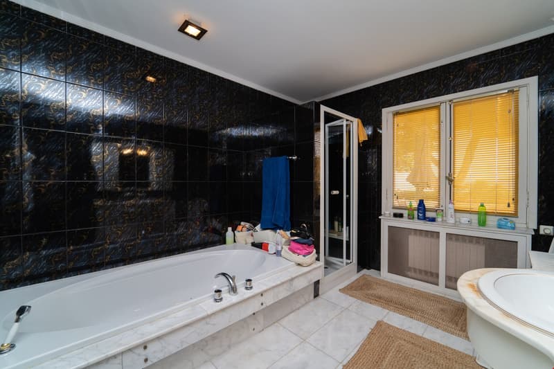 Large bathroom with black and gold patterned tiles covering the walls, a white bathtub, a vanity with a mirror, and a window with yellow blinds.