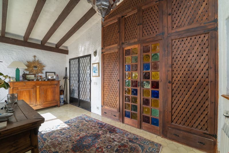 Rustic living room with wooden beams, ornate wooden cabinets with colorful glass inserts, a large ornate mirror, and a patterned area rug on the tile floor.