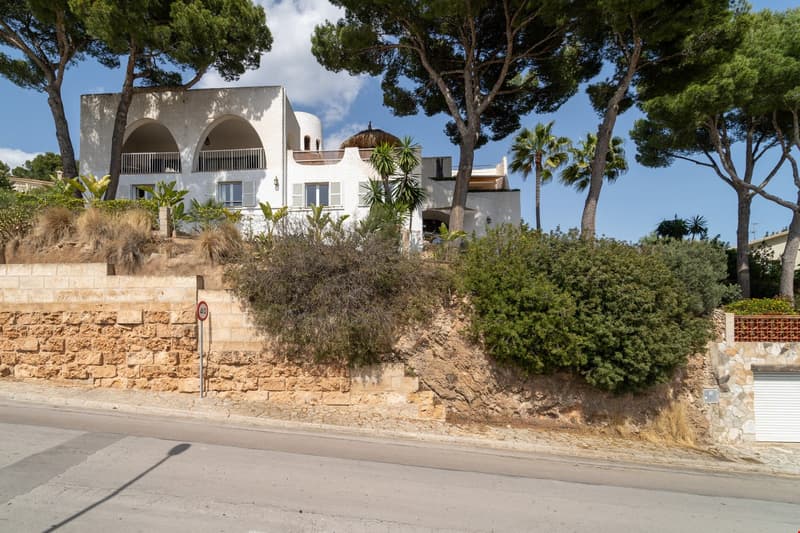 Two-story white Mediterranean-style villa with arched windows and balconies, surrounded by lush greenery and palm trees on a hillside location.