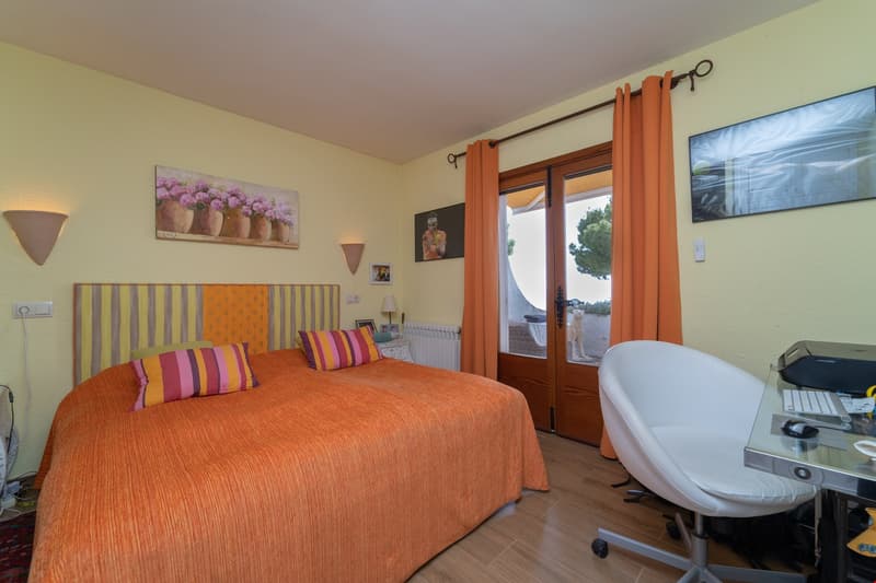 Spacious bedroom with a large bed, colorful striped bedding, wooden furniture, and a balcony or terrace visible through the window. The room has a warm, cozy atmosphere with yellow walls and decorative artwork on the walls.
