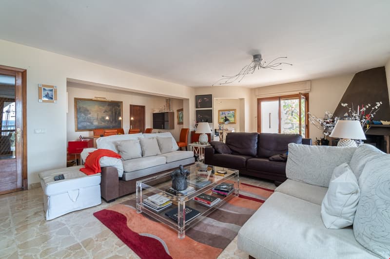 Spacious living room with hardwood floors, beige walls, large windows, gray and brown sofas, glass coffee table, artwork on the walls, and a modern chandelier lighting fixture.