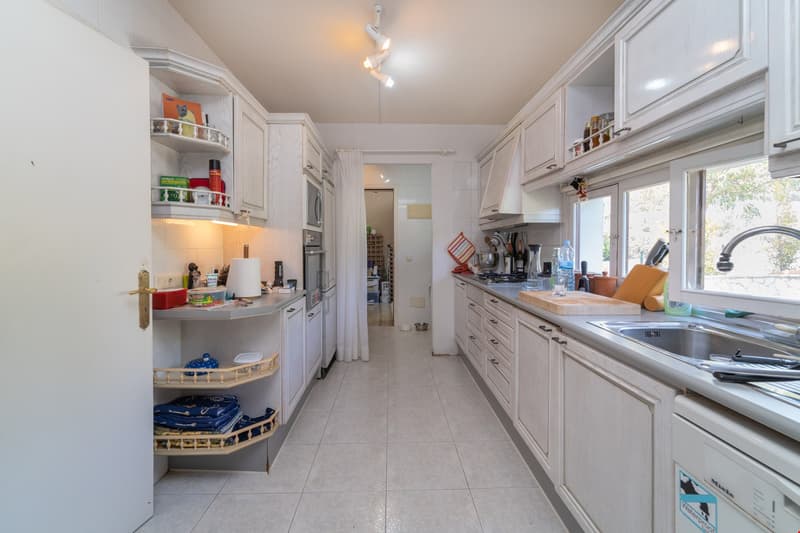 Spacious galley-style kitchen with white cabinetry, tile flooring, and various kitchen appliances and utensils visible. The kitchen includes a washing machine and dishwasher.
