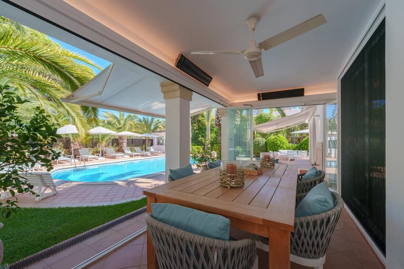 Covered patio with wooden dining table and chairs, overlooking a swimming pool surrounded by palm trees and lush greenery