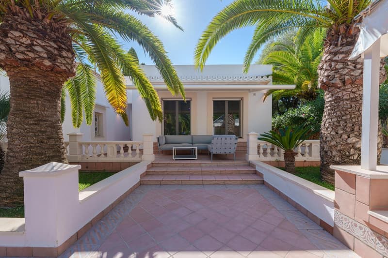 Two-story Mediterranean-style villa with a covered patio, palm trees, and a tiled walkway leading to the entrance.