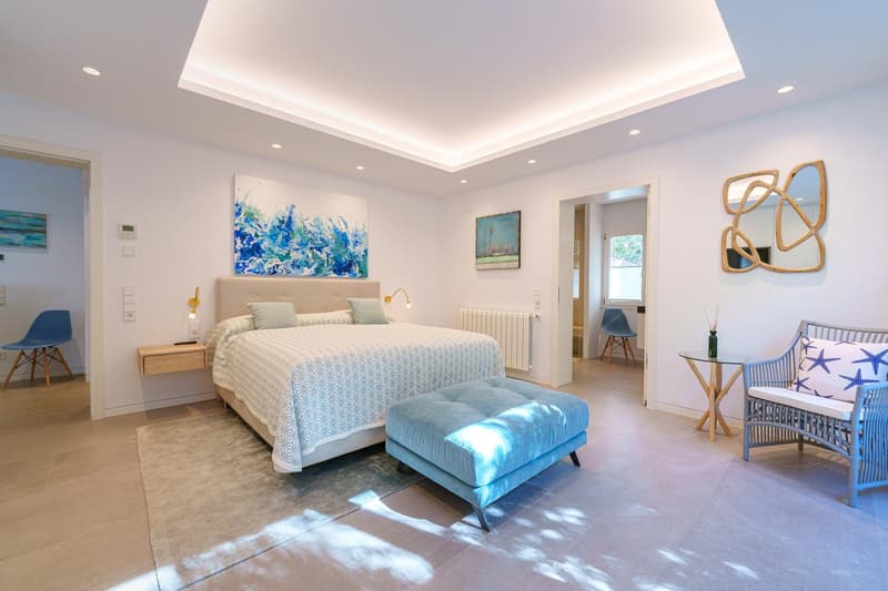 Spacious bedroom with a large bed, blue and white patterned bedding, and a blue velvet ottoman. The room features white walls, recessed lighting, and abstract artwork. There is a balcony or terrace visible through the window.