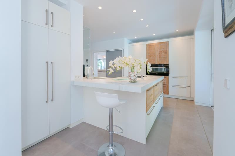 Spacious modern kitchen with white cabinets, marble countertops, built-in appliances, and a central island with bar seating. The kitchen features a dishwasher and is decorated with a vase of white orchids.