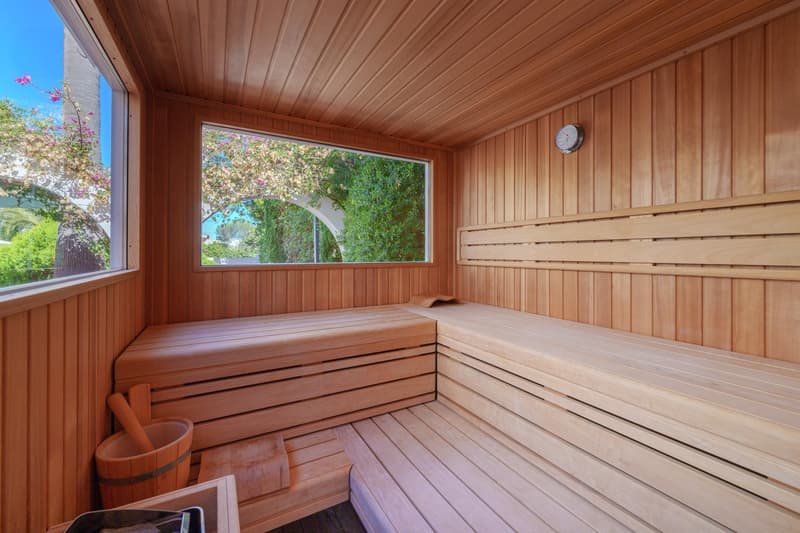A cozy wooden sauna room with a large window overlooking the lush greenery outside. The room features wooden benches and a clock on the wall, creating a relaxing and tranquil atmosphere.