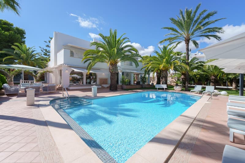 This image depicts a luxurious villa with a large swimming pool surrounded by palm trees and lush greenery. The villa has a modern, white exterior with balconies and terraces. The pool area features comfortable lounge chairs and umbrellas, creating an inv