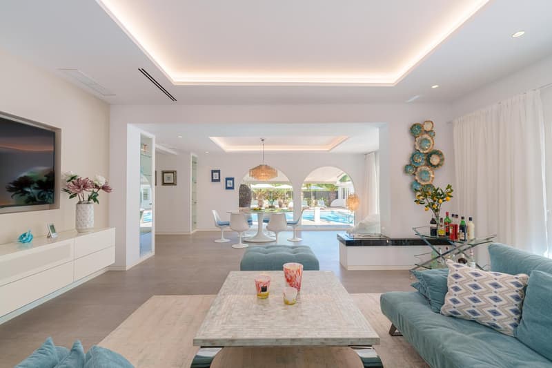 Spacious open-concept living area with large windows overlooking a pool, modern furniture including a sectional sofa and tulip dining table, decorative wall art, and a wet bar area
