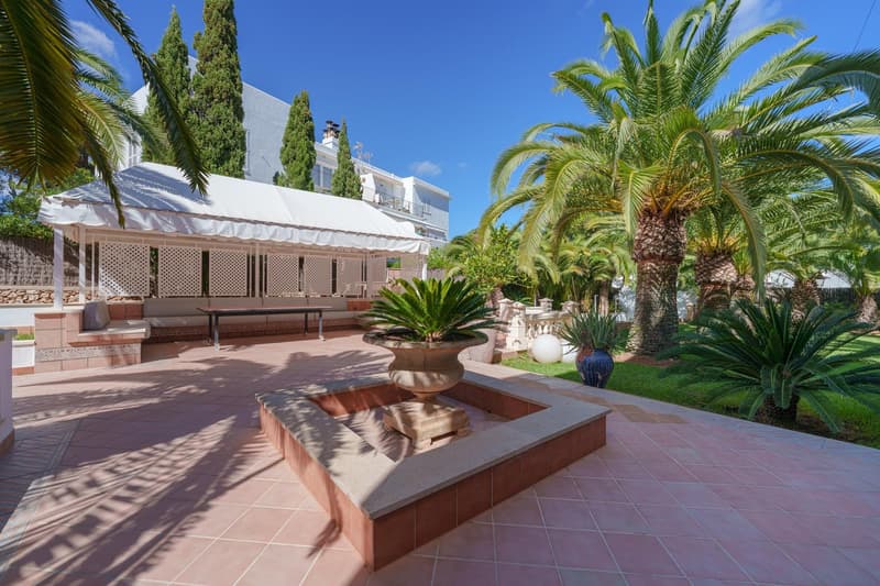 Covered patio with seating area, surrounded by lush palm trees and greenery, with a swimming pool in the foreground and a multi-story building in the background.