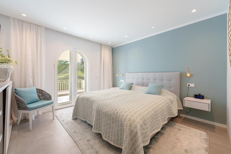 Spacious bedroom with large arched windows leading to a balcony, gray and blue color scheme, tufted headboard, patterned bedspread, and a wicker chair
