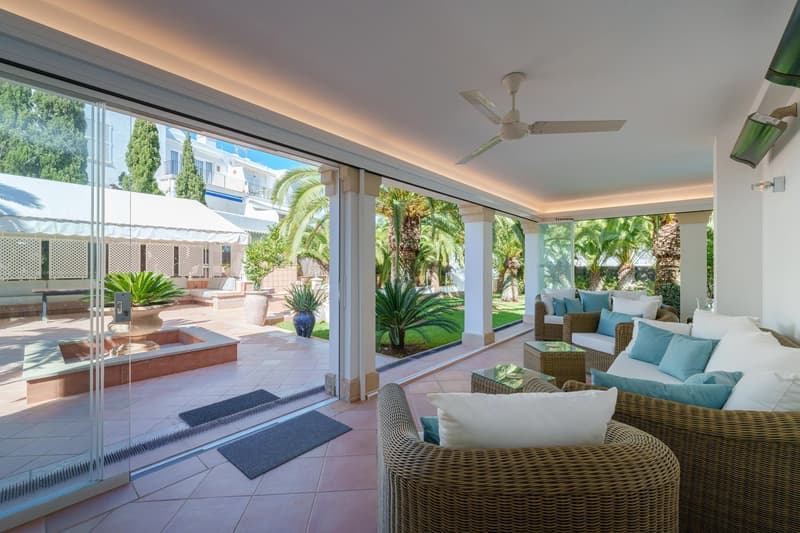 Spacious living room with large windows overlooking a lush garden and swimming pool, wicker furniture, tiled flooring, ceiling fan