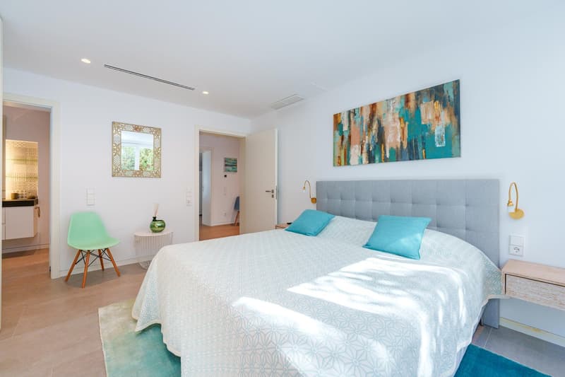 Spacious bedroom with white walls, large windows, gray upholstered headboard, white bedding, abstract art painting, and a green accent chair.