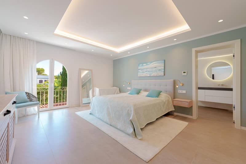 Spacious bedroom with large arched windows, plush white bedding, tufted headboard, recessed lighting, and access to a balcony or terrace overlooking a garden area.