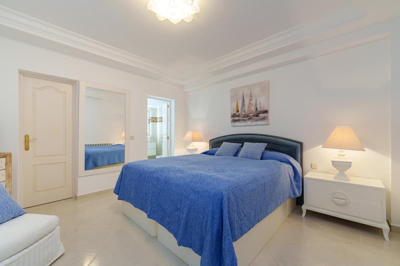 Spacious bedroom with a large bed, blue bedding, white walls, and a chandelier. There is a painting on the wall and a nightstand with a lamp on each side of the bed.