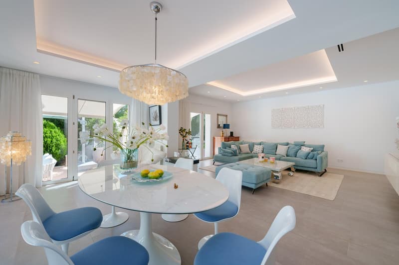 Spacious open-plan living and dining area with large windows, a crystal chandelier, a round white marble dining table with blue chairs, a plush blue sectional sofa, and a decorative wall art piece. The room has a modern and elegant design with neutral col