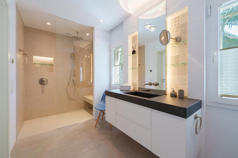 Spacious bathroom with a large walk-in shower, modern vanity with a black countertop, and decorative wall tiles. The room has ample natural light from the window and recessed lighting.