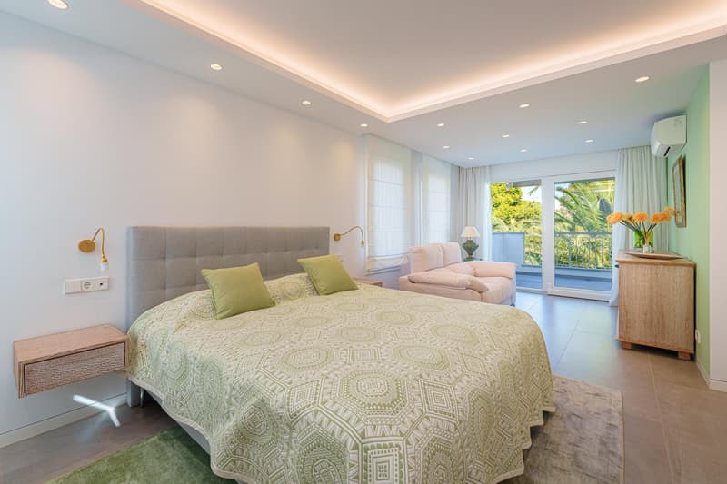 Spacious bedroom with a large bed, gray upholstered headboard, green and white patterned bedding, nightstands, and a seating area with a sofa overlooking a balcony or terrace with a view of greenery outside.