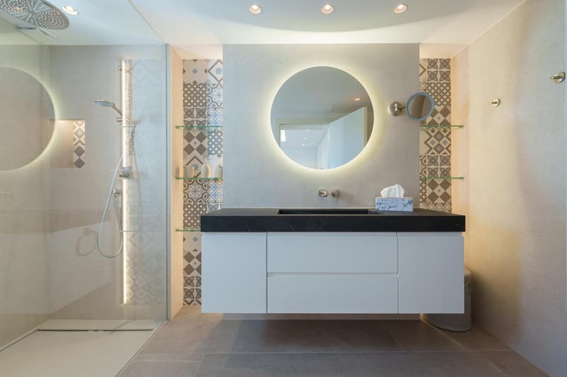 Modern bathroom with a large circular mirror, white vanity with black countertop, tiled walls with a patterned design, and a glass shower enclosure.