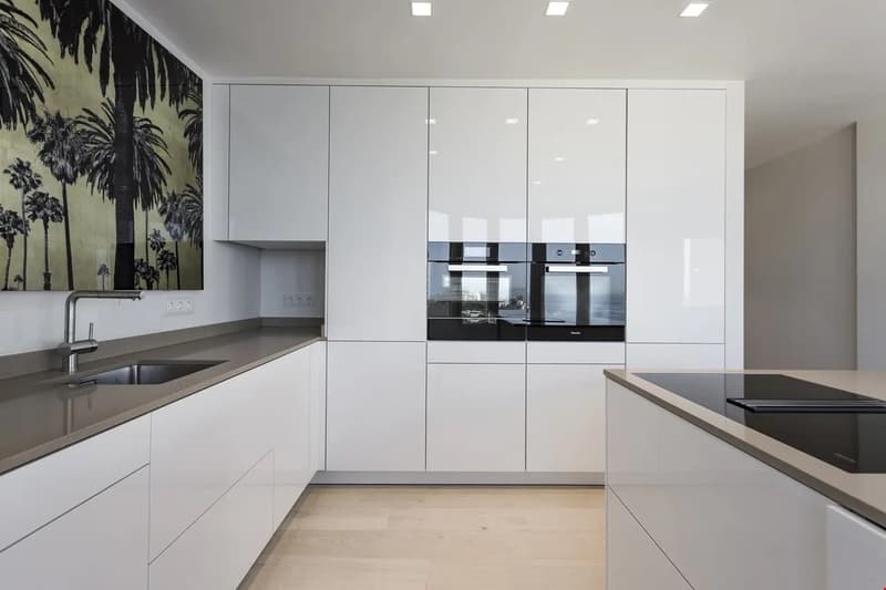 Modern, minimalist kitchen with white cabinets, built-in appliances including a dishwasher, and a large mural of palm trees on the wall.