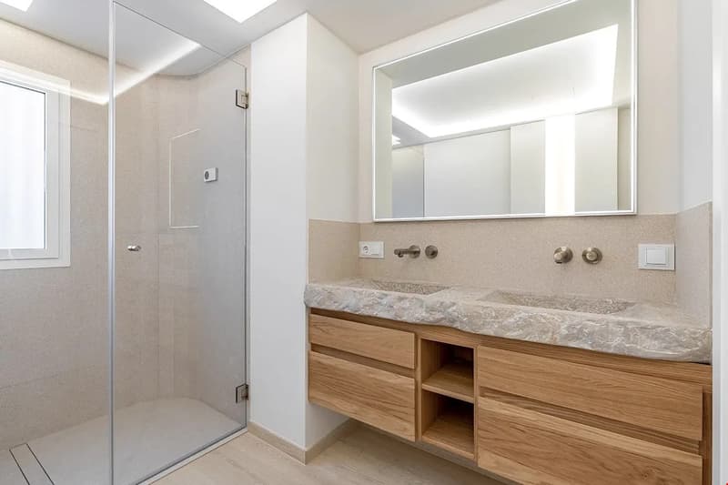 Spacious bathroom with a large vanity cabinet made of wood, a marble countertop, and a walk-in shower with glass enclosure. The room has a modern and minimalist design with neutral colors and ample natural lighting.