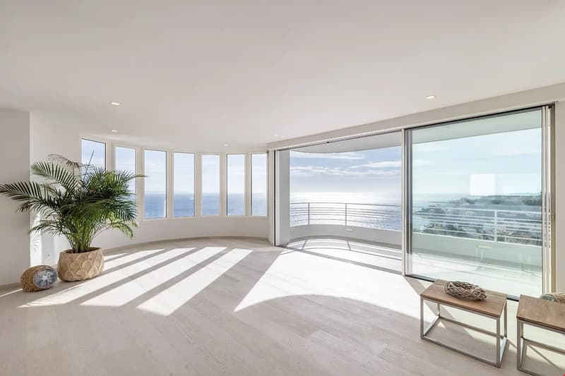 Spacious living room with large windows offering a panoramic view of the ocean. The room has a minimalist and modern design with light-colored hardwood floors, white walls, and a palm tree plant. There are sliding glass doors leading to a balcony or terra