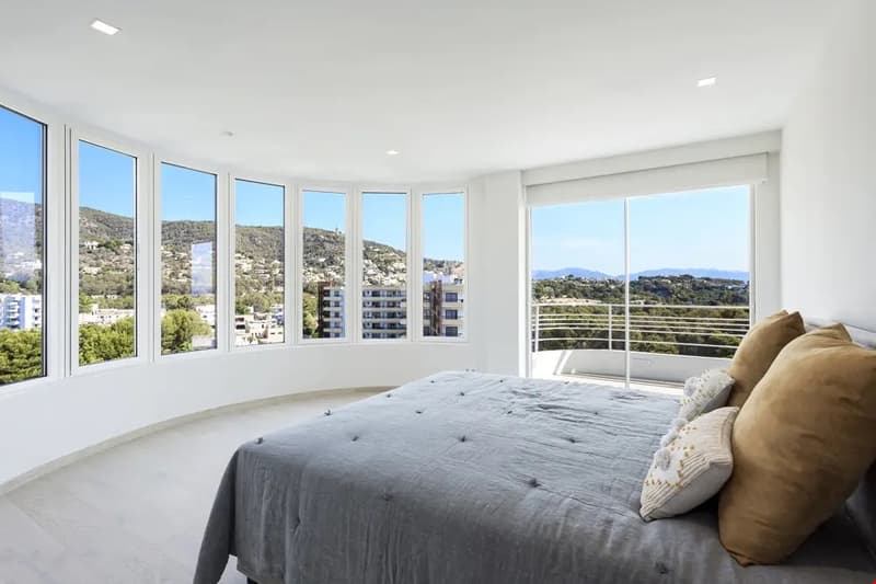 Spacious bedroom with large windows offering panoramic views of the surrounding hills and cityscape, hardwood floors, grey bedding, and a cozy seating area by the windows