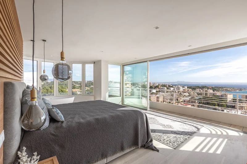 Spacious bedroom with floor-to-ceiling windows offering a panoramic view of the city and the sea. The room features a large bed with a gray comforter, pendant lighting, and a modern, minimalist design.