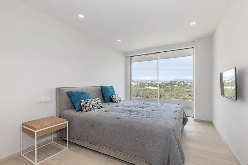 Spacious bedroom with large windows offering a scenic view, grey upholstered bed with colorful pillows, wooden side table, and a flat-screen TV mounted on the wall.