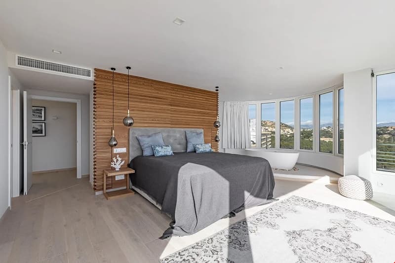 Spacious bedroom with large windows offering a scenic view, wooden accent wall, grey bedding, pendant lighting, and hardwood flooring.