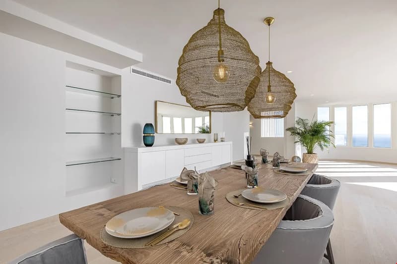 Spacious dining room with a large wooden dining table, gray upholstered chairs, and two large pendant light fixtures with a woven design. The room has white walls, built-in shelving, and large windows providing natural light. The decor includes various de