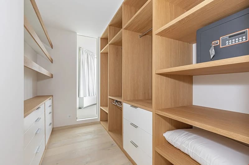 Spacious walk-in closet with wooden shelves and drawers, a safe, and a mirror. The room has a minimalist design with white walls and wooden accents.