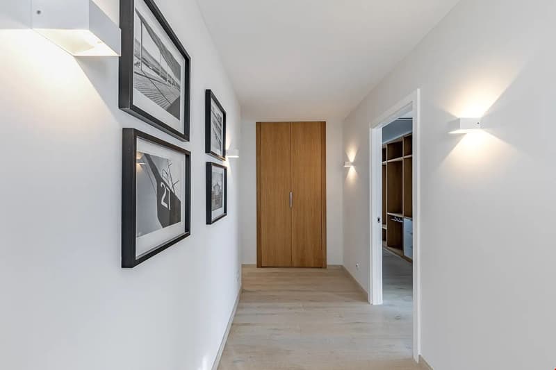 Spacious hallway with white walls, hardwood flooring, and a wooden cabinet door. The hallway features framed artwork on the walls and recessed lighting.