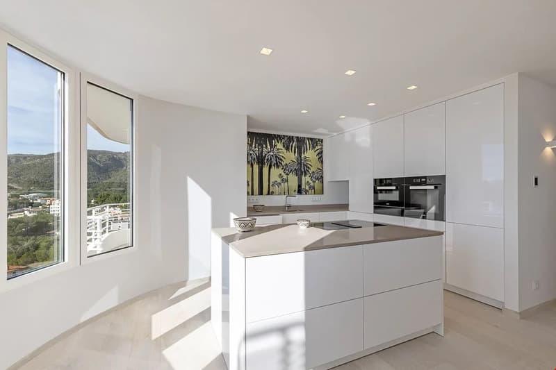 Spacious modern kitchen with white cabinets, quartz countertops, and high-end appliances. Large windows provide a scenic view of the surrounding hills and landscape.