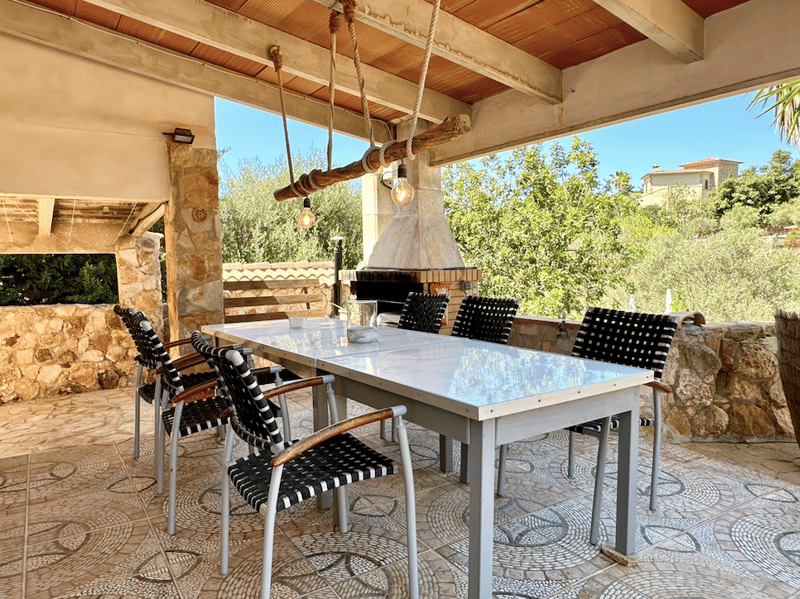 Covered outdoor dining area with a large stone fireplace, wooden beams, and hanging light fixtures. The dining table has a glass top and is surrounded by black and white striped chairs. The area overlooks a lush, green garden.