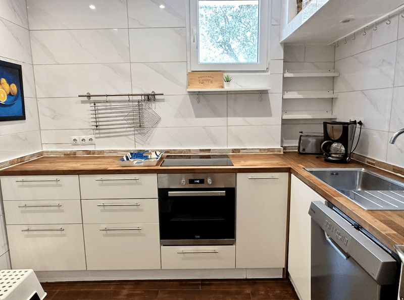Modern kitchen with white cabinets, wooden countertops, built-in oven, dishwasher, and shelves for storage. Large window overlooking greenery outside.