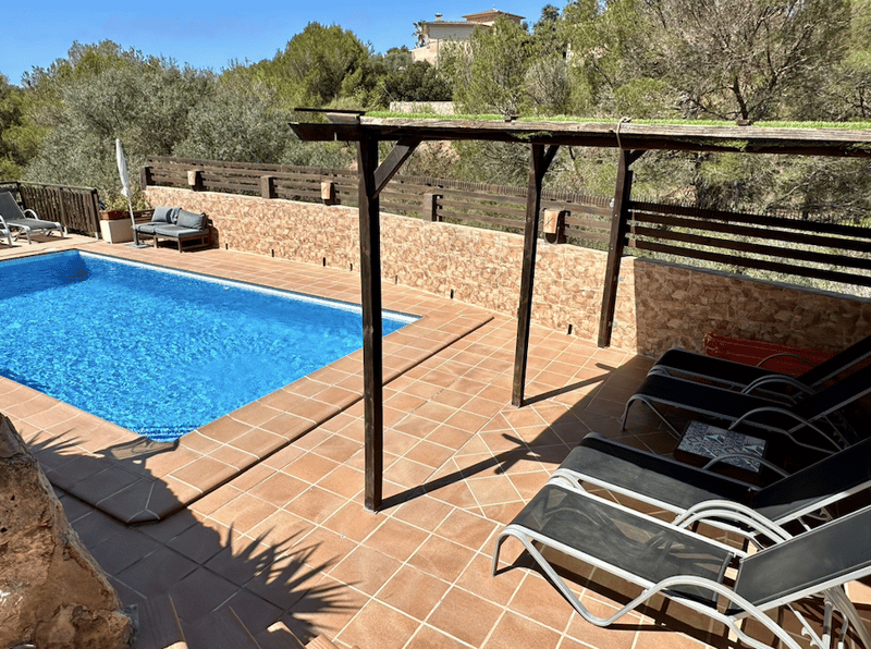 Outdoor pool area with a wooden deck, stone walls, and a covered seating area. The pool is surrounded by lounge chairs and has a view of the surrounding trees and buildings.