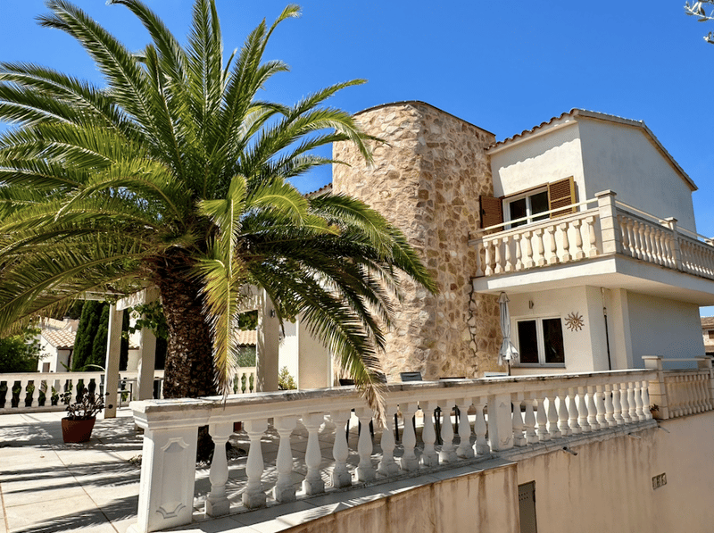 Two-story Mediterranean-style villa with stone exterior, white walls, and a balcony. There is a palm tree in the foreground.