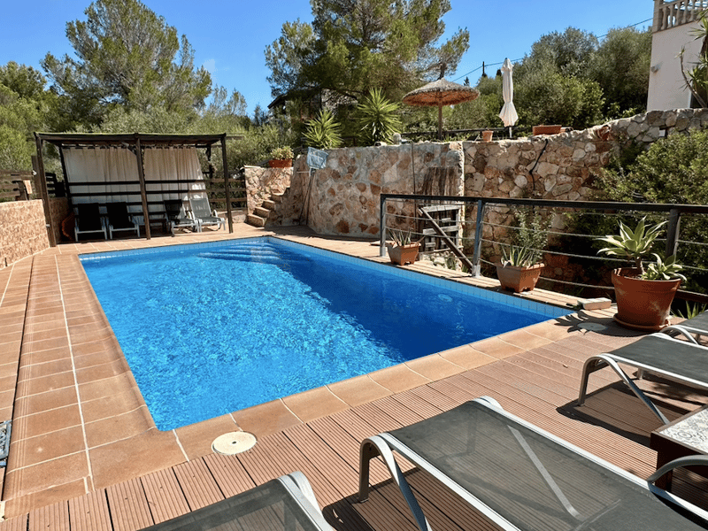 Outdoor swimming pool with wooden deck, surrounded by stone walls, palm trees, and patio furniture. There is a covered patio area with an umbrella.