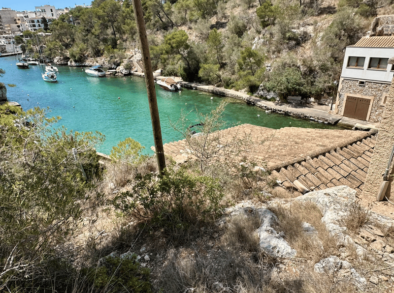 The image shows a scenic coastal landscape with a turquoise blue bay or inlet surrounded by lush vegetation and rocky cliffs. There are several boats moored in the calm waters. A building with a tiled roof can be seen on the hillside, suggesting a residen