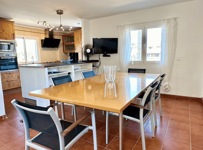 The image shows an open-concept kitchen and dining area. The kitchen features wooden cabinets, a large countertop, and a built-in dishwasher. The dining area has a rectangular wooden table with chairs, and a flat-screen TV mounted on the wall. The room ha