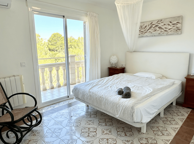 Spacious bedroom with large windows, balcony, white bedding, wooden furniture, and patterned tile flooring