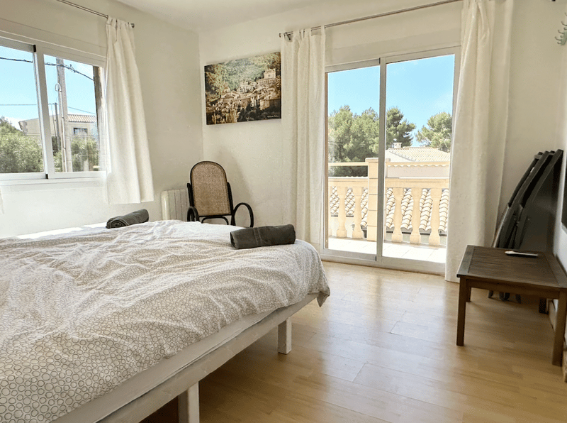Spacious bedroom with large windows, hardwood floors, a bed with patterned bedding, a chair, and a balcony overlooking the outdoors.