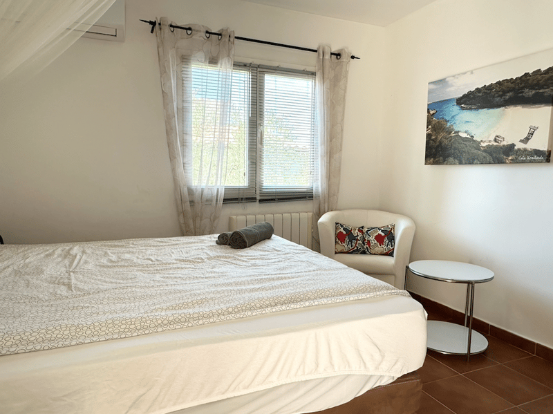 Spacious bedroom with large windows, white walls, hardwood floor, king-size bed, armchair, and a framed artwork on the wall depicting a scenic coastal landscape.