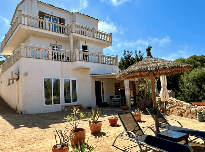 2-story Mediterranean-style villa with balconies, white exterior, tiled roof, patio with lounge chairs and a thatched-roof gazebo, potted plants and landscaping