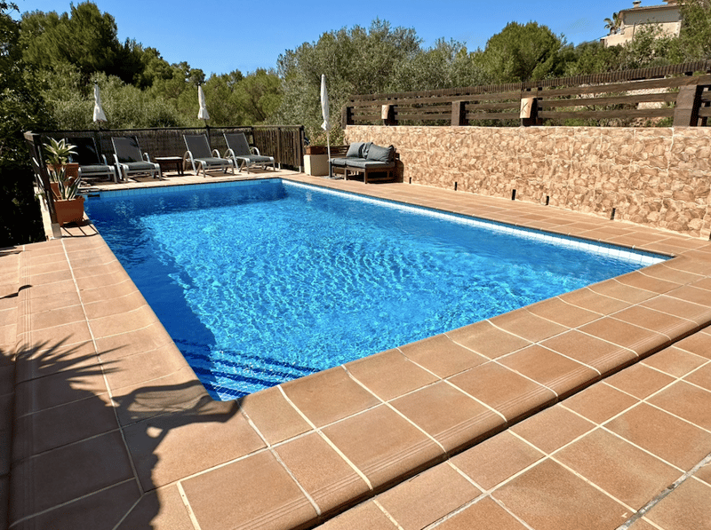 Rectangular outdoor swimming pool with clear blue water, surrounded by a stone wall and wooden deck with lounge chairs, in a lush, green setting with trees and shrubs.