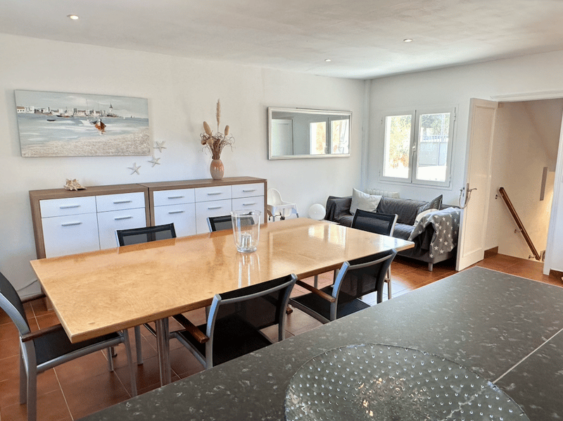 The image shows an open-plan living and dining area. The living room has a gray sofa and a decorative mirror on the wall. The dining area has a large wooden table with black chairs. There is a sideboard with drawers and a vase of flowers. The room has lar