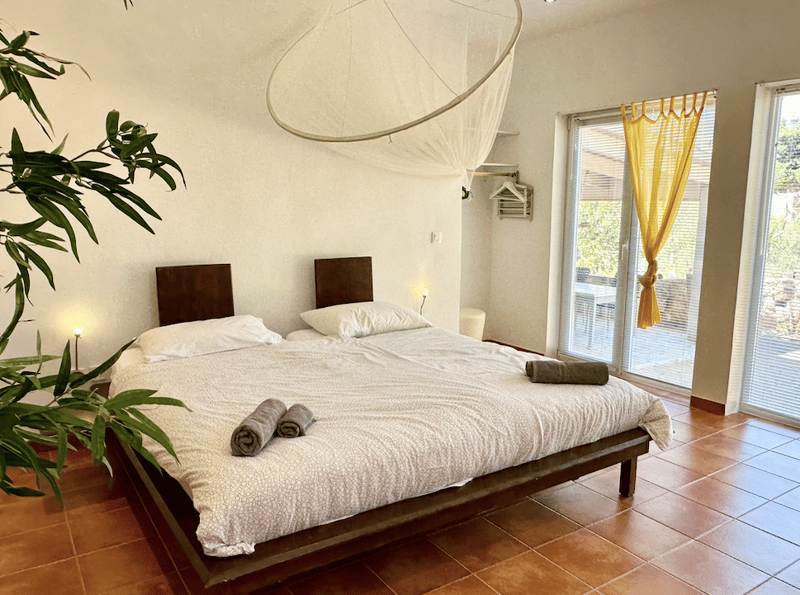 Spacious bedroom with a large bed, wooden headboards, and a sliding glass door leading to a balcony or terrace. The room has a neutral color scheme with white bedding and a hanging light fixture. There are also some potted plants adding a natural touch to